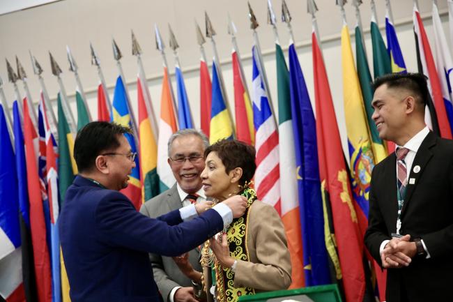 A representative presents a traditional necklace to Sheam Satkuru, ITTO Executive Director, as a gesture of appreciation and cultural honor, during the ITTC60 session, with colorful national flags in the background.