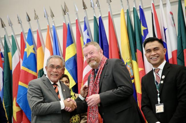 Two delegates exchange a warm handshake during the ITTC60 session, with one presenting a traditional scarf as a token of appreciation, while colorful national flags create a vibrant backdrop.