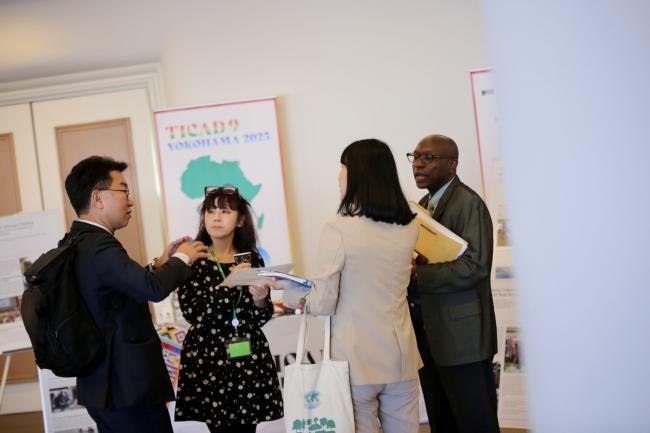 Delegates networking at ITTC-60, with a TICAD9 Yokohama 2025 poster in the background.