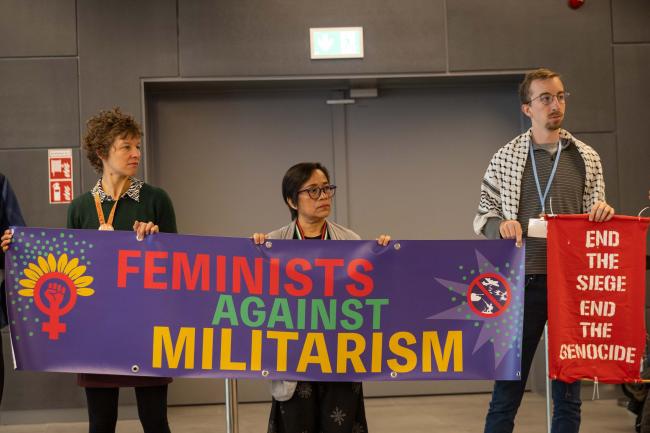 Members of civil society demonstrate in the corridors, calling to 'end genocide everywhere', highlighting the links between links between militarism, military emissions, military spending, climate injustice, environmental destruction, displacement, and gender inequality
